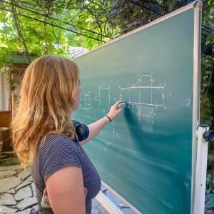 Caro Harwell '24 stands in front of a black board and traces a finger along a diagram drawn on the board. Harwell wears a black and white striped shirt and has long blond hair. 绿色的树叶在头顶和背景中.