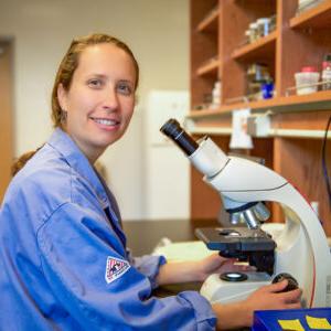 Tessa Solomon-Lane sits in front of a microscope in a laboratory while smiling at the camera. She has long dark blond hair pulled back and wears a blue lab coat.