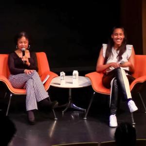 Anna Malaika Tubbs and Jan Barker Alexander sit side by side in orange chairs on a black stage. Barker Alexander holds up a mic while speaking to the audience.